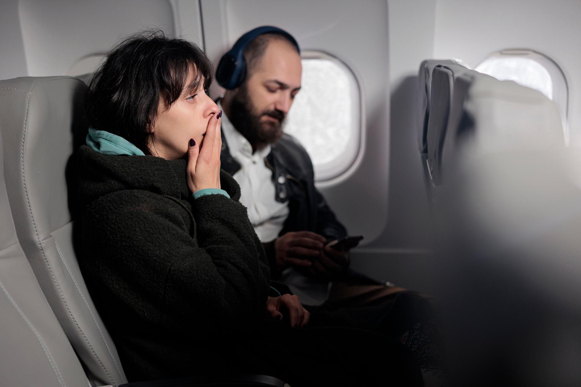 Femme angoissée en avion, mettant en lumière l'anxiété liée à l'aviophobie, tandis que son voisin reste calme, illustrant les différentes réactions face à la peur de l'avion.