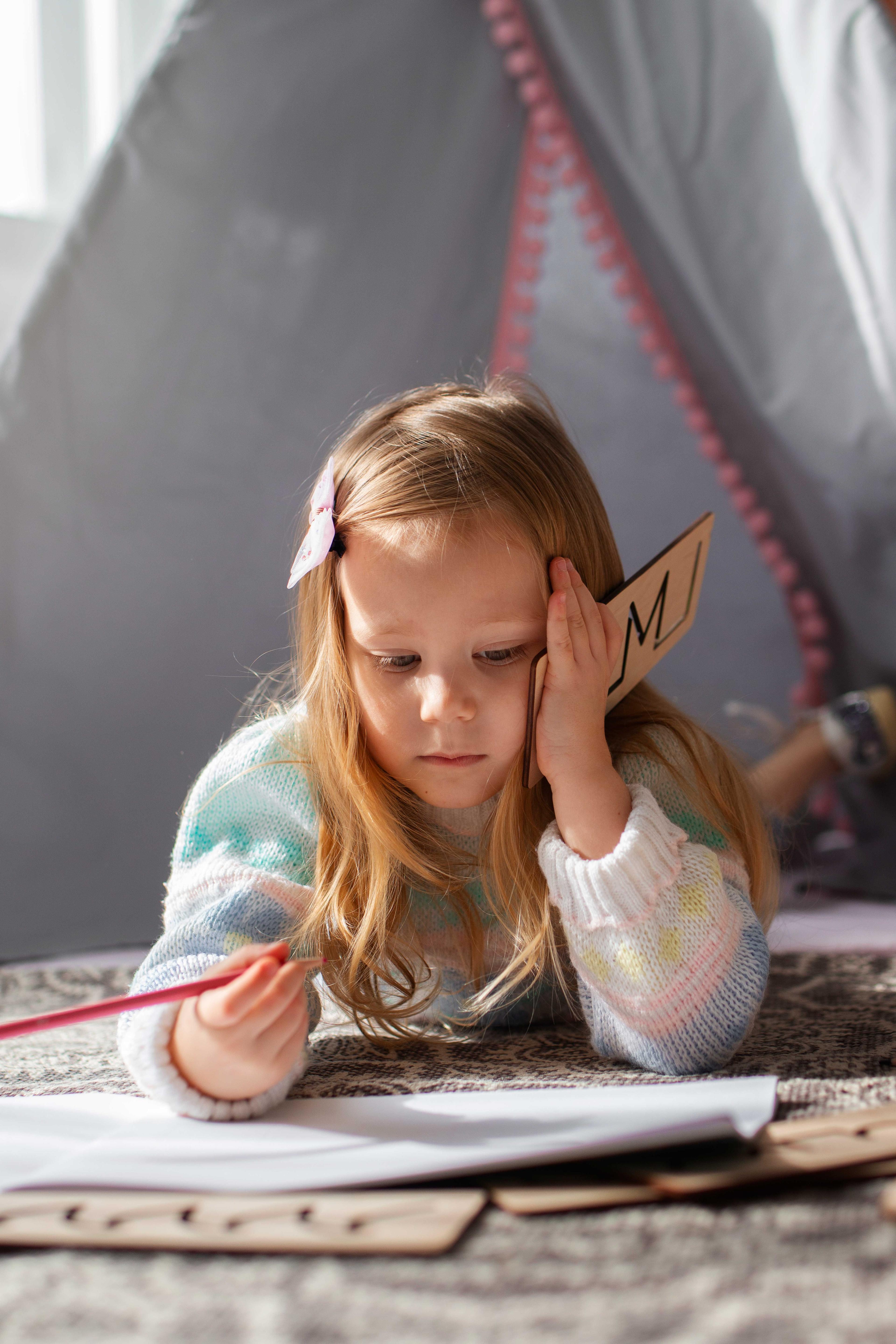 Fillette allongée, concentrée sur son cahier, tenant une lettre en bois, illustrant les difficultés d’apprentissage liées à la dyslexie.