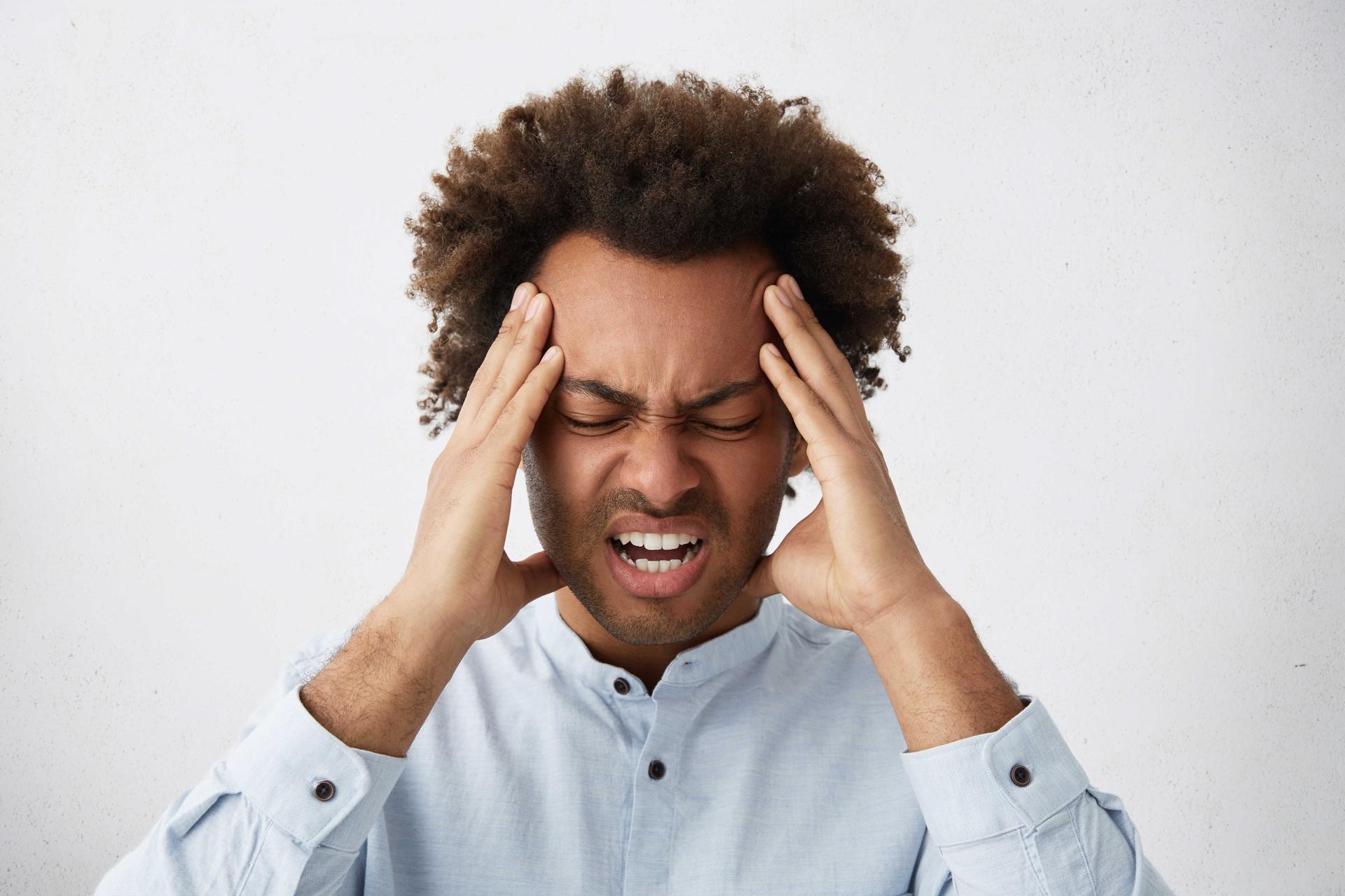 Homme, les yeux fermés, tenant sa tête des deux mains, exprimant une douleur ou une tension liée au stress. L'arrière-plan est clair, soulignant le focus sur son expression tendue et accablée.